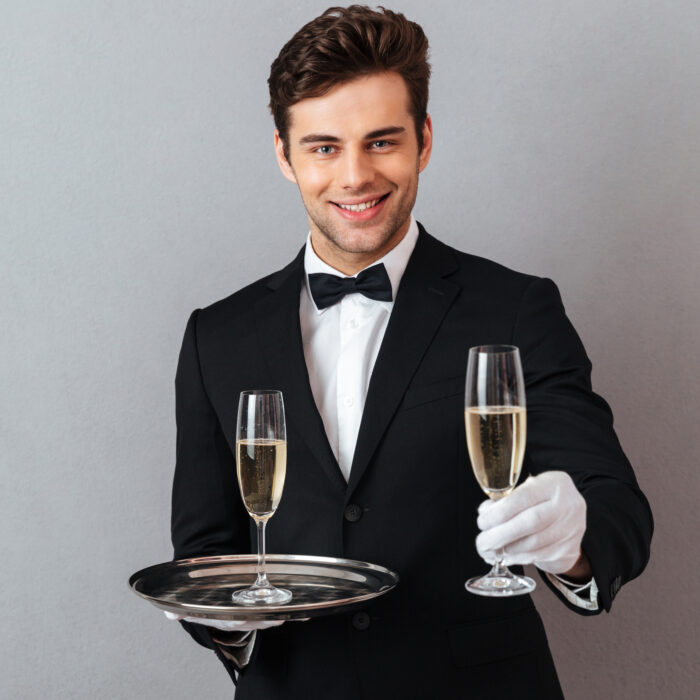 Photo of cheerful young waiter standing isolated over grey wall background. Looking camera holding glass of champagne give it to you.