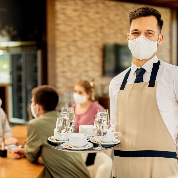 Happy waiter serving coffee to his customers while wearing protective face mask in a cafe due to coronavirus epidemic.